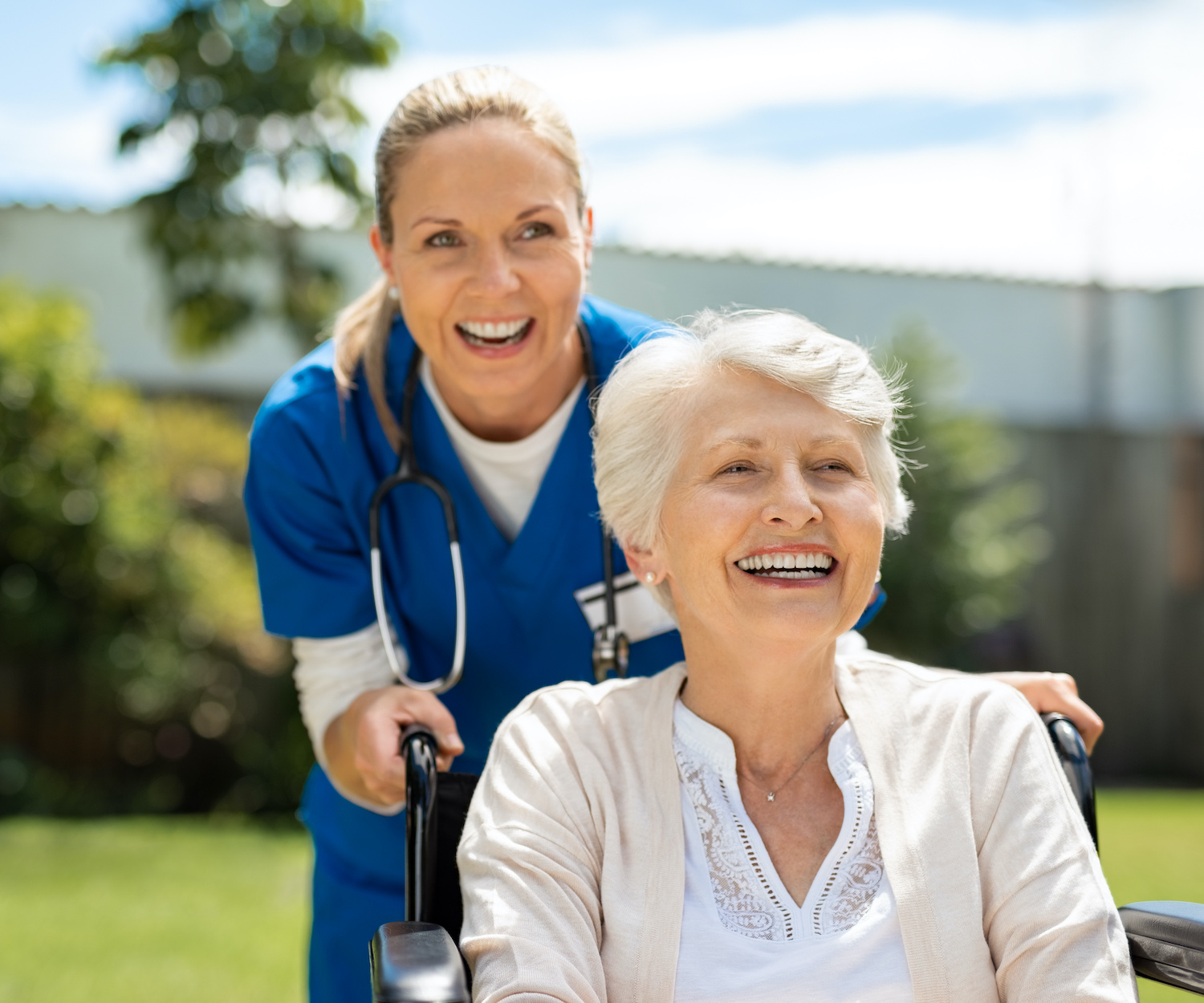 Happy nurse and elderly woman sitting in wheelchair enjoying outdoor treatment session. Beautiful nurse with laughing senior woman in wheelchair at outdoor park. Smiling disabled old lady in wheelchair at park.
